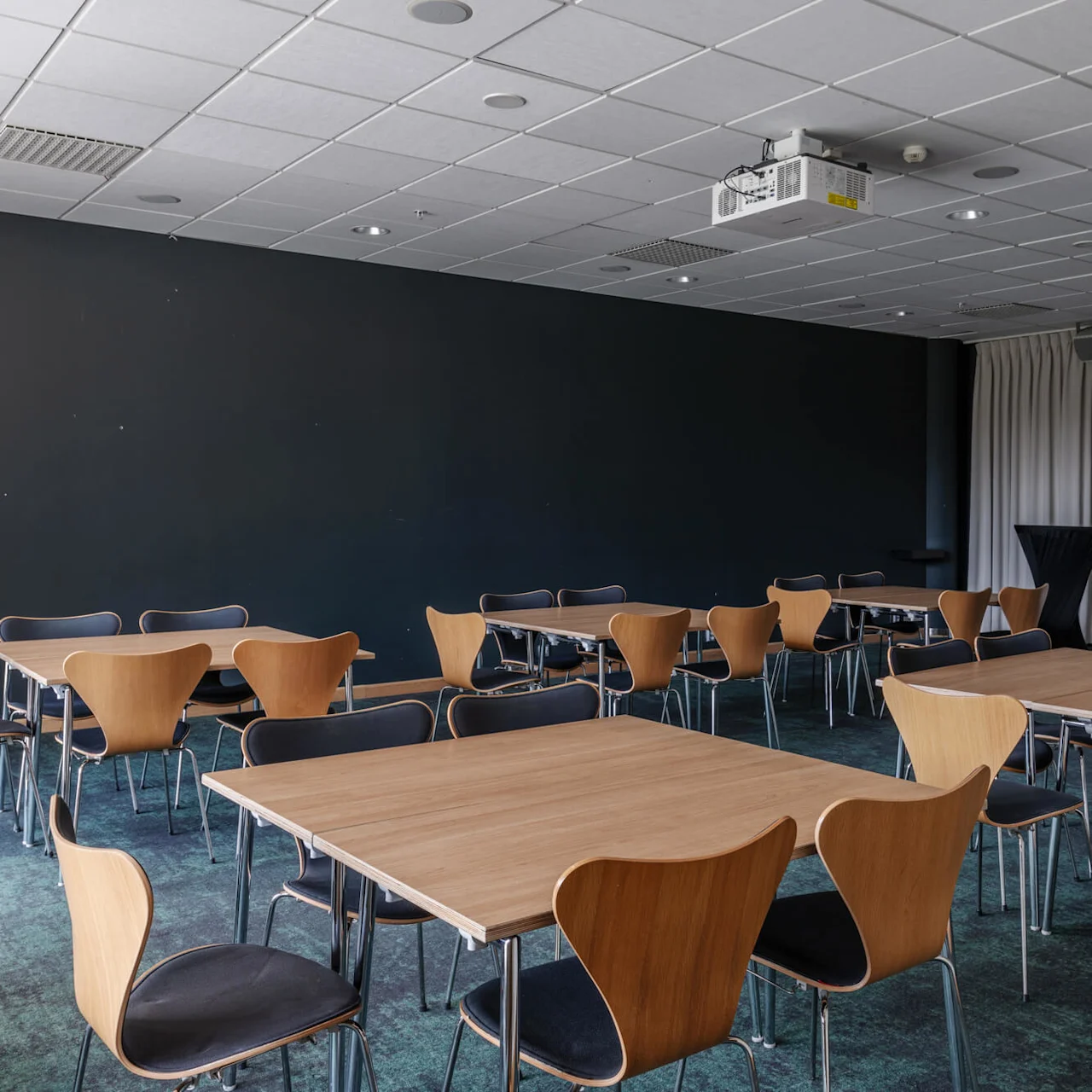 Bord och stolar placerade i ett mötesrum med en takmonterad projektor, mörka väggar och dragna gardiner, vilket antyder en konferens- eller presentationsmiljö.