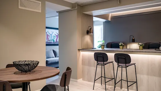 Round wooden table with black chairs sits near a modern kitchen counter featuring high stools; decorative plants and artwork adorn the cozy, softly-lit open-plan living area.
