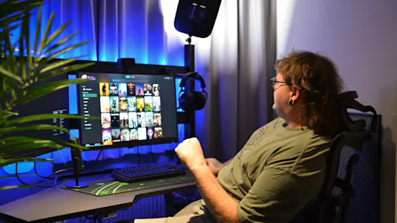 En person sidder ved et skrivebord og ser på en computerskærm med medieminiaturer. Rummet er svagt oplyst med blåt lys, og planter og gardiner omgiver opsætningen.