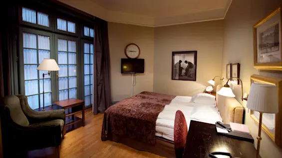 Et godt opplyst hotellrom med en elegant oppredd seng, et sitteområde, en veggmontert TV og innrammede kunstverk, utstråler en koselig og sofistikert atmosfære.