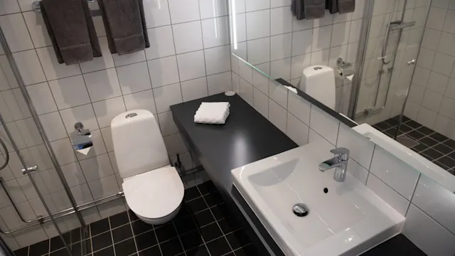 Toilet and sink are installed in a tiled bathroom. The toilet is adjacent to a counter with a folded towel. A large mirror and shower area enhance the space.