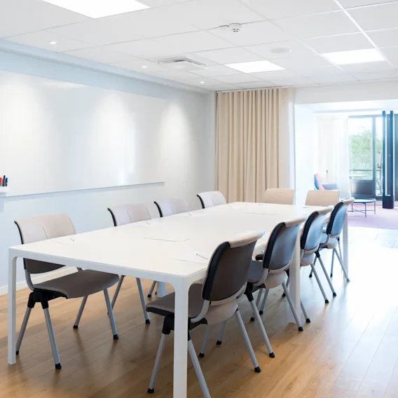 Ett modernt konferensrum med ett långt vitt bord omgivet av stolar, bredvid ett stort fönster. Väggarna har en whiteboard; en klädhängare håller plagg. Dörrmärke lyder "Terassen 2."