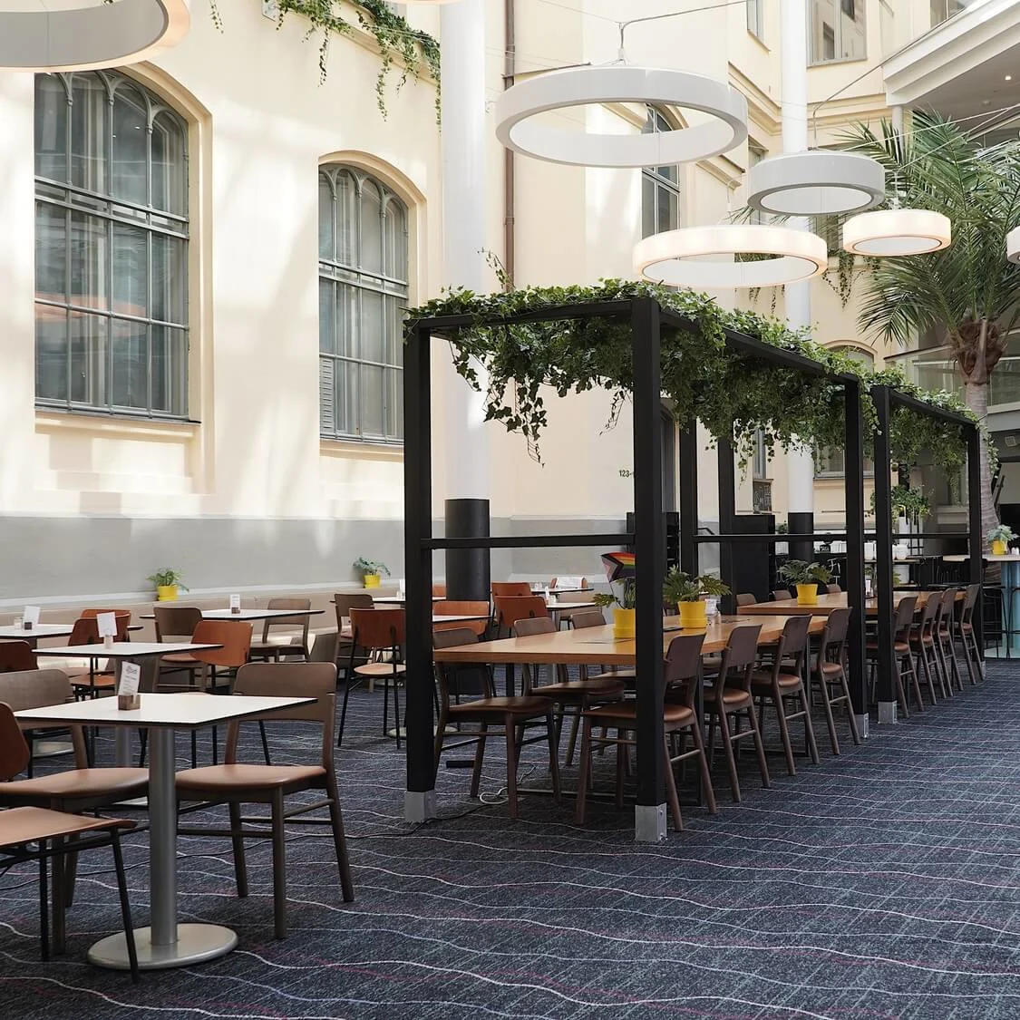 An indoor atrium cafe with tables, chairs, and a bar counter, sunlight streaming through arched windows, and plants adorning the space.