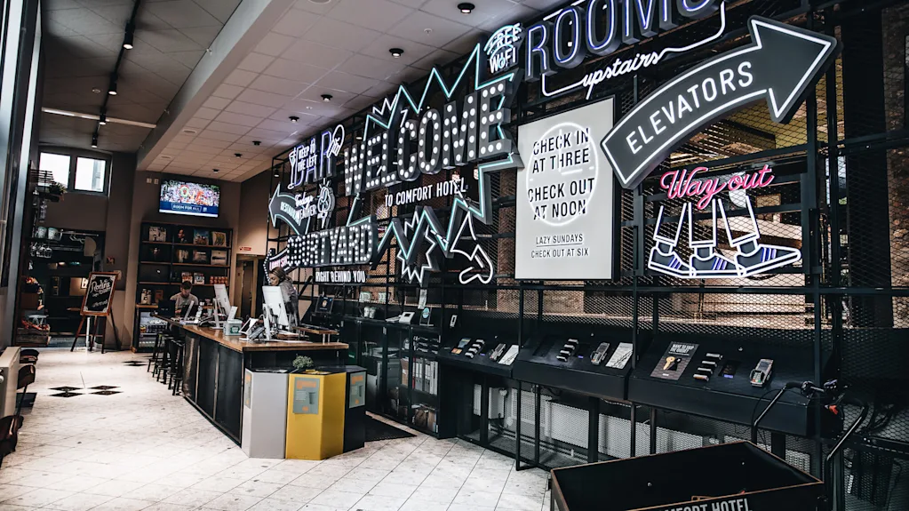 Reception counter with two staff members working, surrounded by neon signs displaying directions and hotel information. Text: "Welcome", "Check in at three, check out at noon", "Elevators", "Way out".