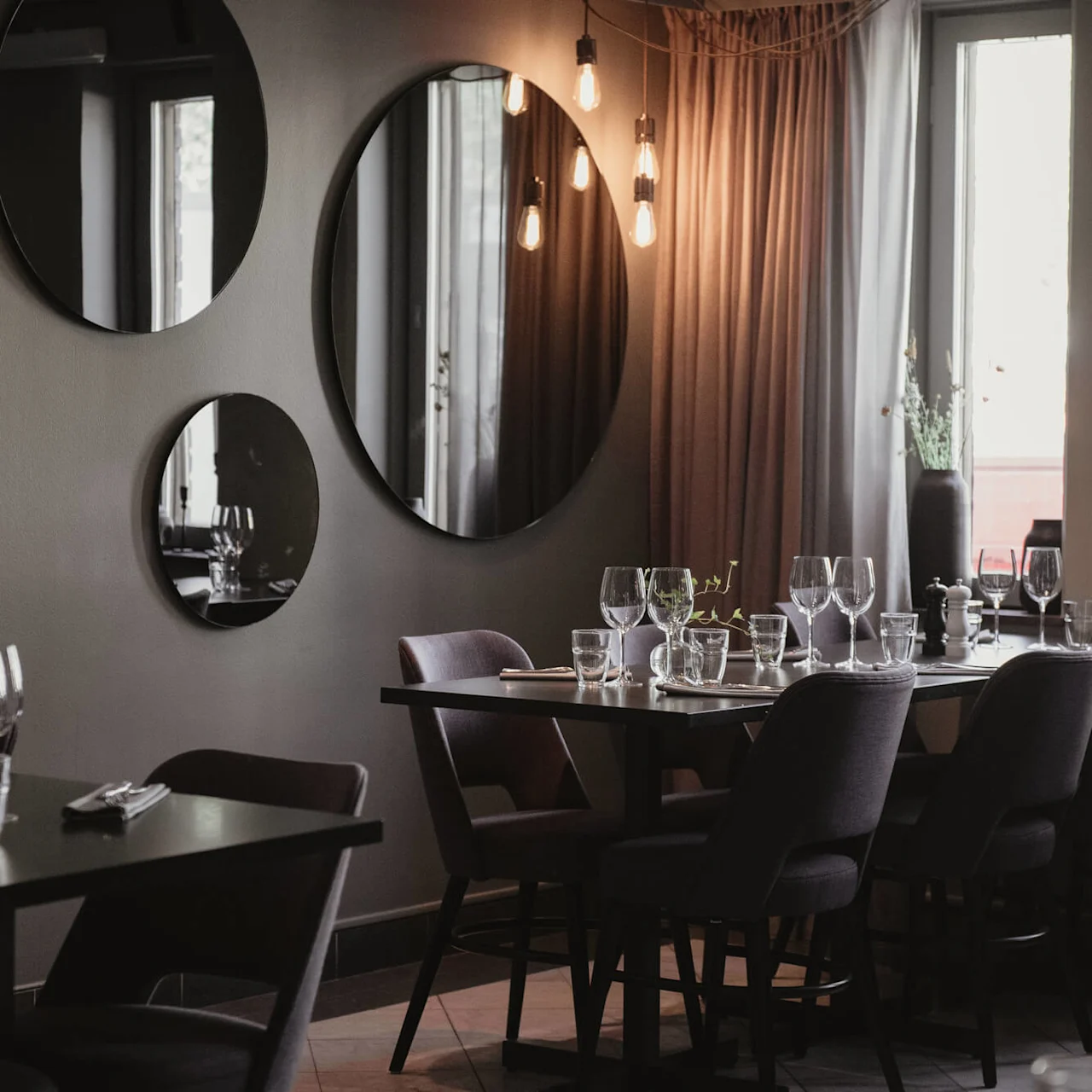 Borden är dukade med vinglas och bestick i en svagt upplyst restaurang. Speglar pryder grå väggar och reflekterar varma hängande lampor. Mjuka gardiner och fönster förstärker den mysiga atmosfären.