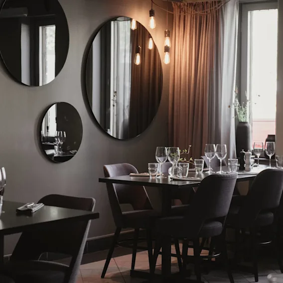 Borden är dukade med vinglas och bestick i en svagt upplyst restaurang. Speglar pryder grå väggar och reflekterar varma hängande lampor. Mjuka gardiner och fönster förstärker den mysiga atmosfären.