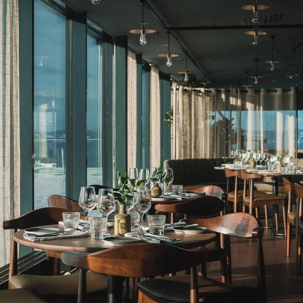 Et stilig restaurantinteriør med dekkede bord, store vinduer og en lys, innbydende atmosfære for bespisning.