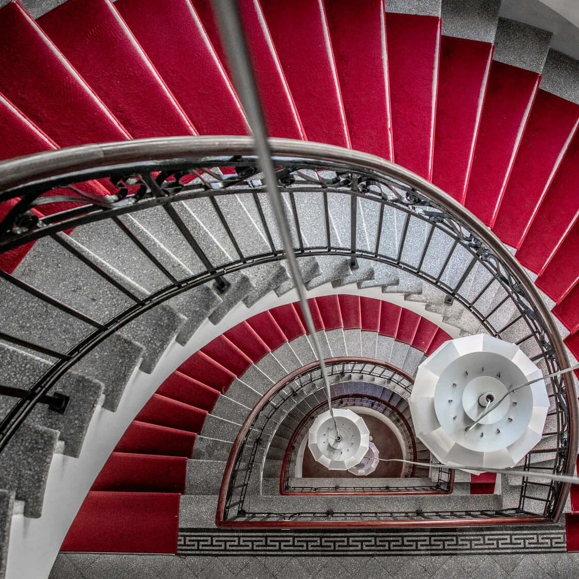 A spiral staircase with red carpeting descends within a building, offering a geometric and architectural perspective from above.