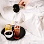 Käsi kaataa cocktailia ravistimesta lasiin valkoisella sängyllä olevalla tarjottimella.