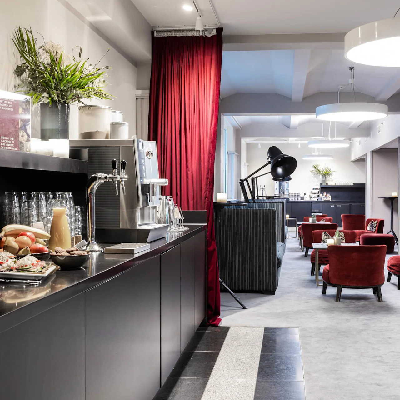Kahvila, jossa on juomia ja välipaloja mustalla tiskillä, ympärillä tyylikkäät istuimet punaisilla tuoleilla modernissa, hyvin valaistussa aulassa. Teksti: "SPECIAL OFFER THE WONDERS OF HOTELNESS".