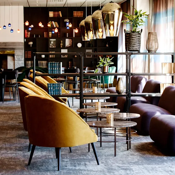 Yellow chairs face round tables in a cozy lounge, with bookshelves and plants. Purple sofas are positioned by sunlit windows with sheer curtains.