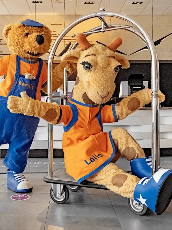 Två kostymklädda figurer, en björn och en giraff, poserar lekfullt på en bagagevagn i en hotellobby. Björnens outfit har texten "Bernie" och giraffens "Lollo".