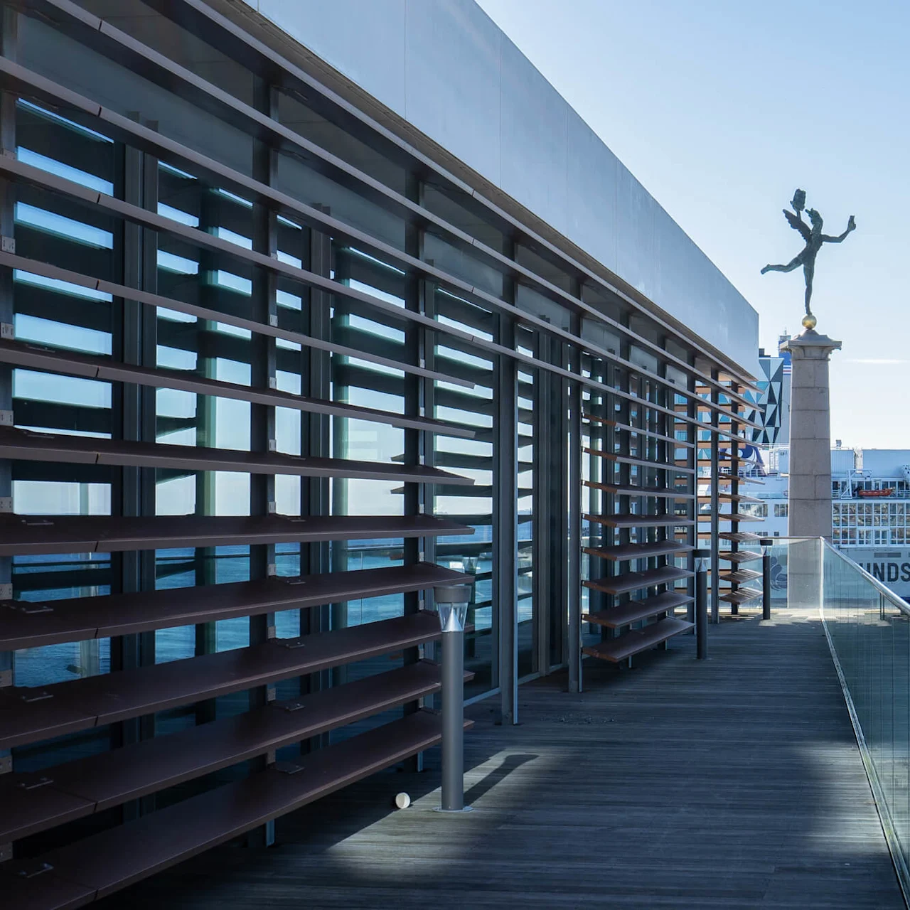 Glassbalkong med utsikt over en havn med en statue på sokkel og en ferge merket "MOLSLINJEN." Moderne bygning med vinklete trelister til venstre. Solrik dag, blå himmel.