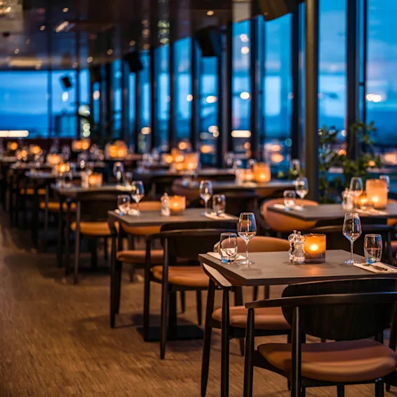 Bordene er dekket med vinglass og lys i en svakt opplyst restaurant, som skaper en elegant atmosfære. Store vinduer har utsikt over en by ved skumring.