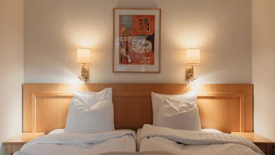 Et fredeligt hotelværelse med to senge, en sengegavl i træ og varme væglamper, der oplyser et indrammet kunstværk ovenfor.