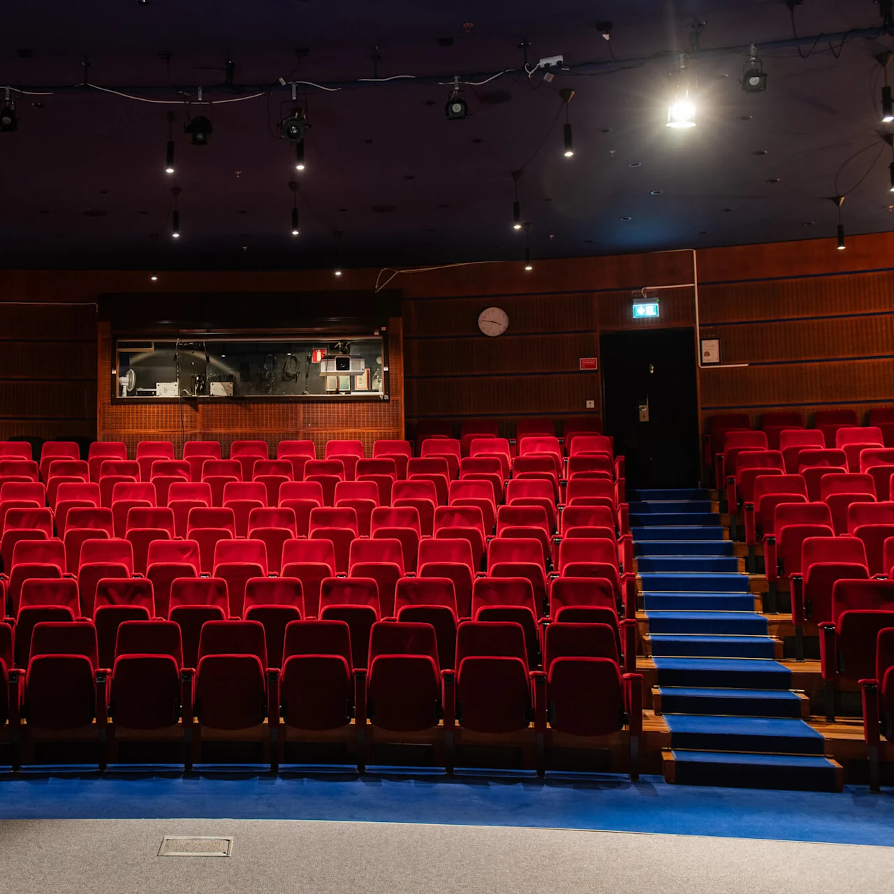 A theater features rows of empty red seats and two central blue-carpeted aisles. Overhead lights illuminate the wooden walls and a control room window. Two “exit” signs are visible.