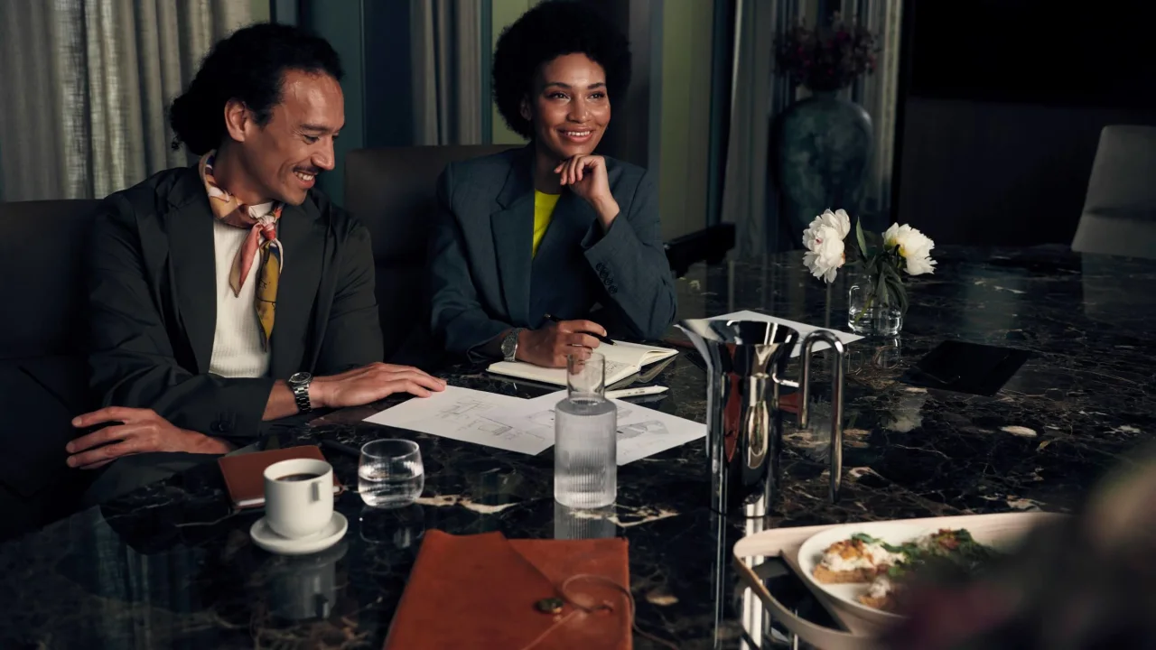 Two professionals, one male and one female, are conversing over documents at a dining table in a dimly lit, upscale room.