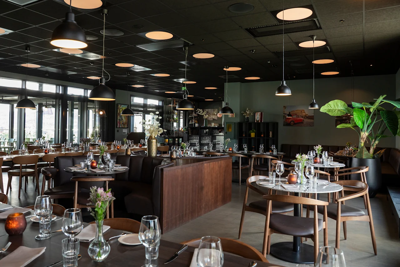Et stilfullt og romslig restaurantinteriør med mange bord dekket for bespisning, med moderne innredning, store vinduer og varm belysning.