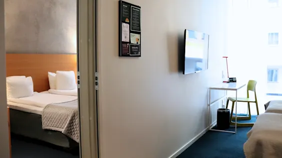 Ljust hotellrum med säng genom dörröppning, TV, skrivbord och anslagstavla: Valkommen, Information, Get started.