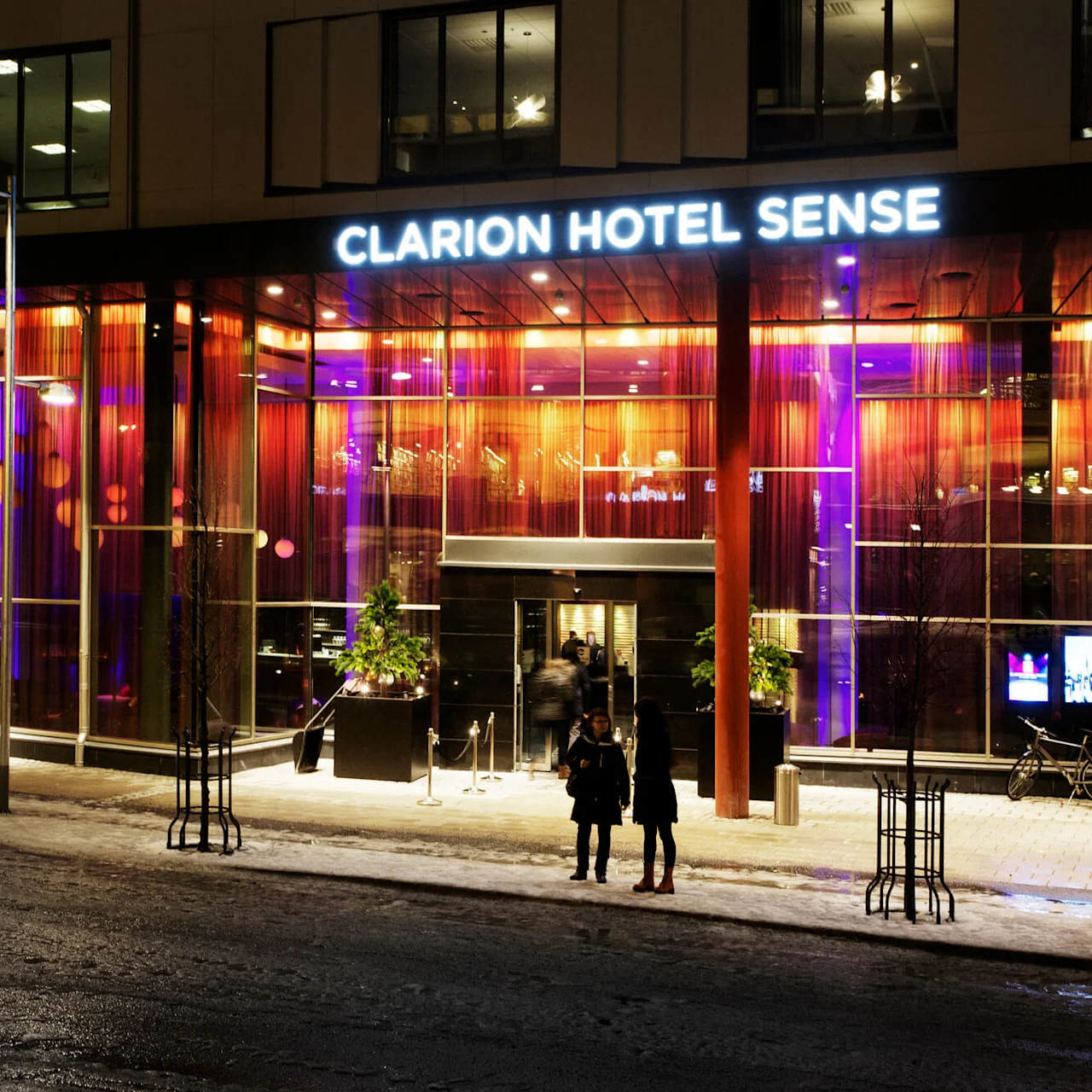 En hotellentré lyser upp på natten, med starka orange och lila ljus genom stora fönster. Två personer står utanför på en snöig väg. Skylten lyder "CLARION HOTEL SENSE".