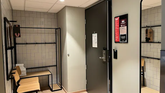 Hotellihuoneen sisäänkäynti tummalla ovella, avoin vaatekaappi, istuimet ja kyltti, jossa lukee DELI, WE CARE, Get benefits.