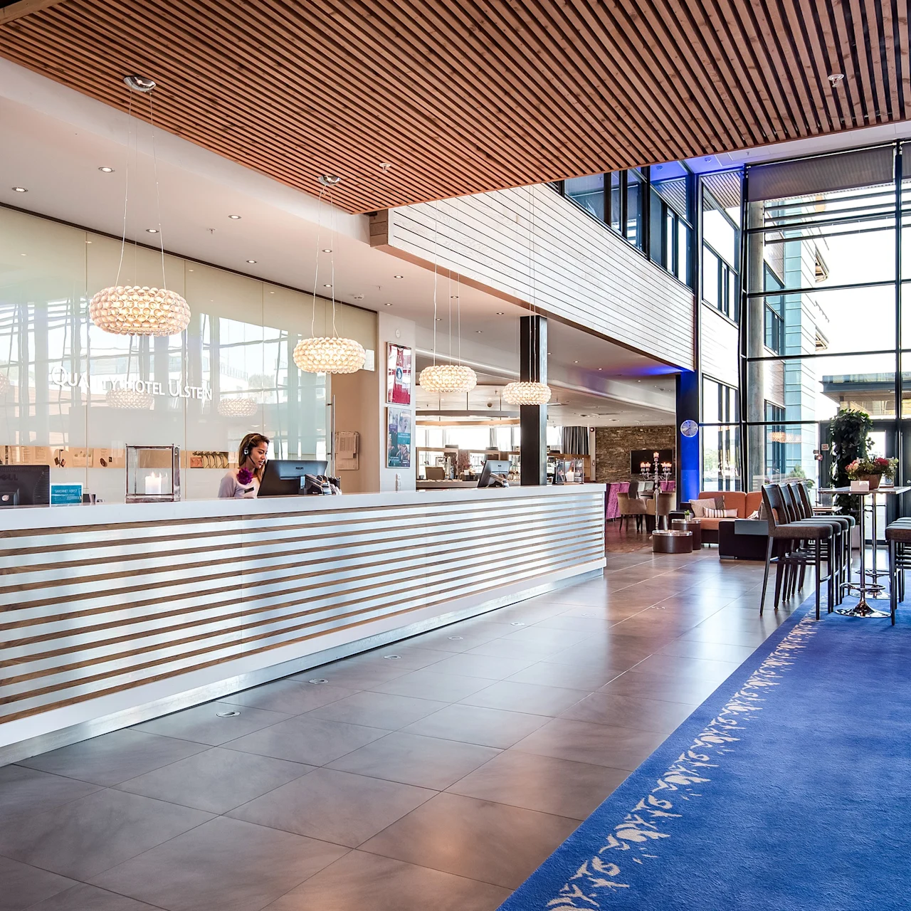 Resepsjonsdisk plassert i en moderne hotellobby, bemannet av en person som arbeider bak den, med stilige lysarmaturer over. Store vinduer fyller området med naturlig lys. Tekst: "QUALITY HOTEL ULSTEIN."