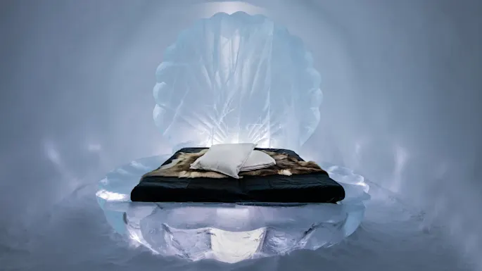 En intrikat utskåret isseng prydet med pels og puter, plassert i et svakt opplyst isrom som ligner et vinterlandskap.