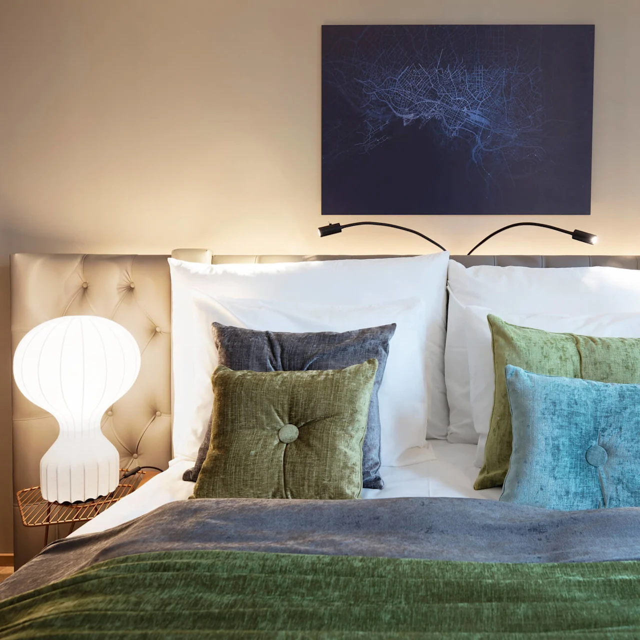 A neatly made bed is adorned with blue and green pillows, flanked by two lit lamps, beneath a dark blue abstract wall art piece in a softly lit bedroom.