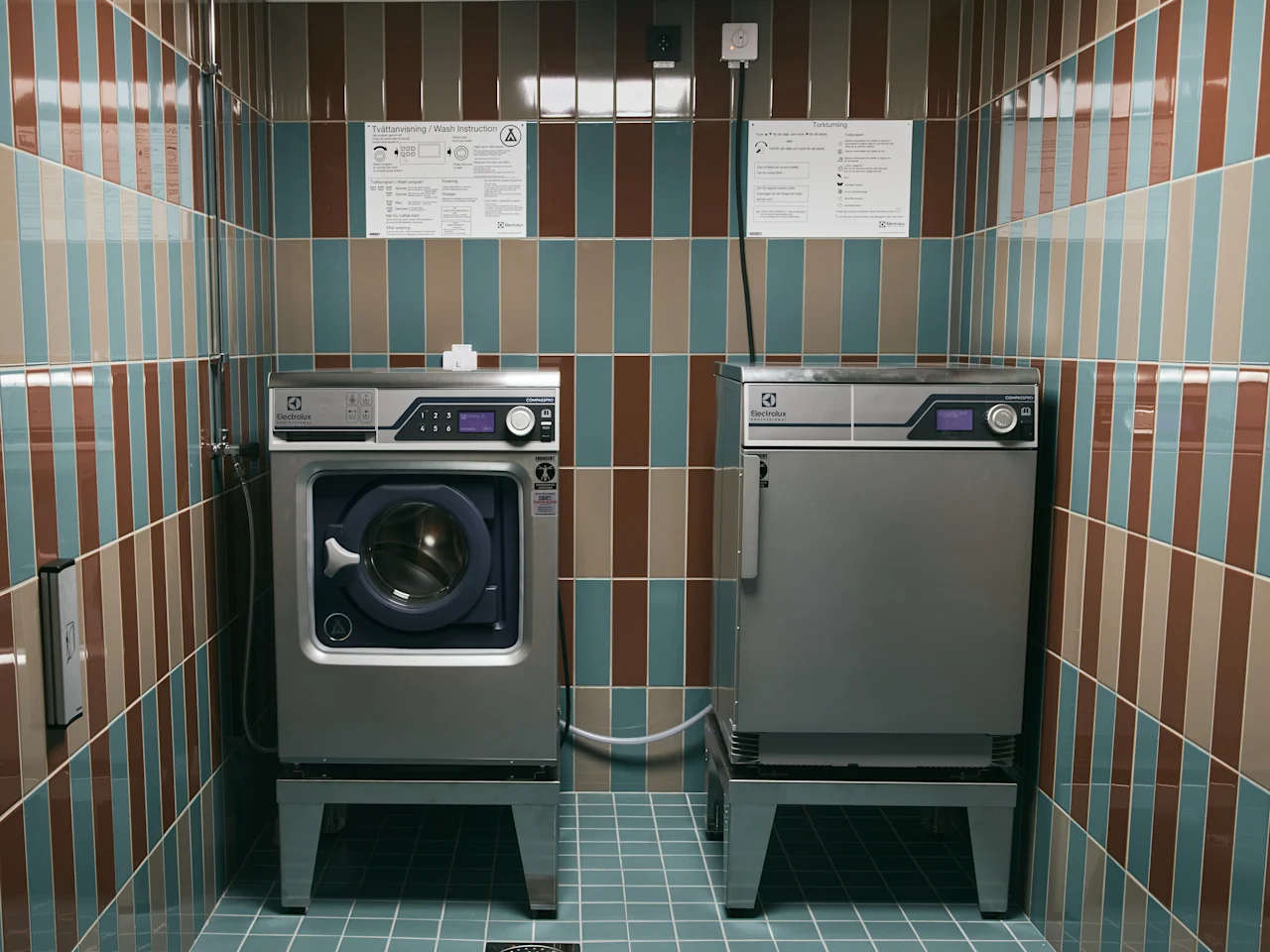 Ett rent tvättstuga med randiga kakel, med en tvättmaskin och torktumlare. Instruktioner för Tvättanvisning och Torkning är uppsatta.