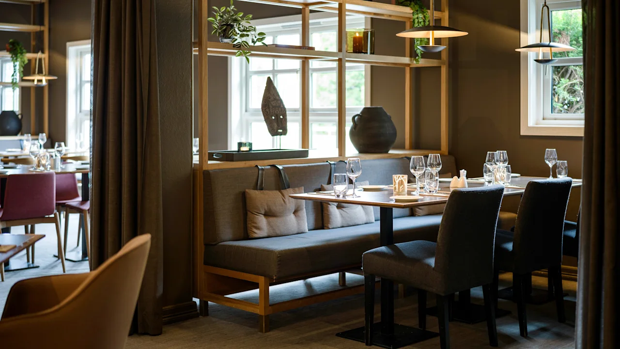 En hyggelig restaurantinteriør med en træskillevæg pyntet med planter og dekorationer. Bordene er elegant dækket med glas og servietter, hvilket skaber en indbydende atmosfære nær store vinduer.