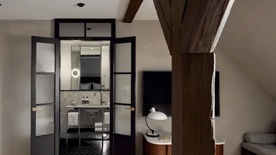 A bathroom with a sink and mirror is seen through open glass doors. Wooden beam, TV, and lamp on a round table in the adjoining room.