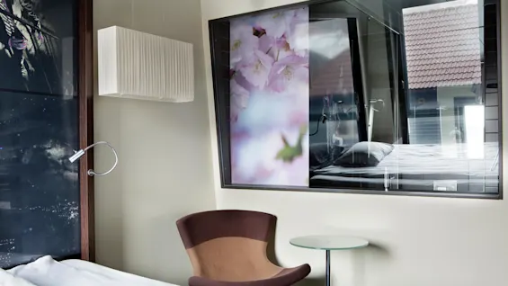En brun stol står ved siden av et glassbord i et moderne rom. Bak en stor blomsterkunstverk og et vindu som reflekterer en seng og ytre hustak.