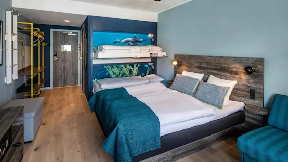 A double bed with teal bedding is centered in a modern hotel room, featuring underwater-themed wall art. A ceiling fan rotates above, and a blue armchair sits nearby.