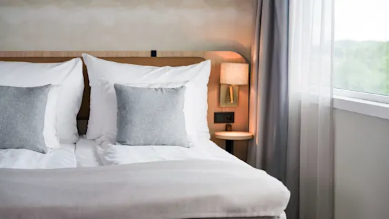 A comfortable hotel room with a neatly made bed, white and grey pillows, a bedside lamp, and a window with natural light.