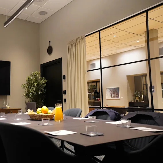 A conference room table holds a bowl of fruit and a glass of orange juice, surrounded by chairs. A large TV on the wall and glass partition complete the setting.
