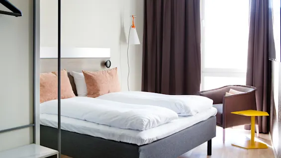 A neatly made bed with white linens and two coral pillows is situated in a modern, brightly lit room. Nearby, a gray armchair and yellow table rest against dark curtains.