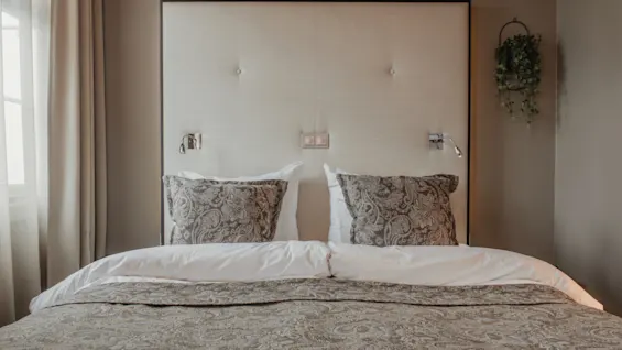 A comfortable hotel room with a neatly made bed, patterned pillows, and wall-mounted lights, suggesting a relaxing stay.