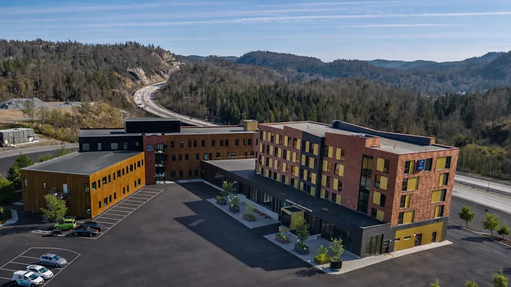 Ett modernt hotellkomplex med parkering, omgivet av skogsklädda kullar och en motorväg i bakgrunden. Synlig text: Hotell.