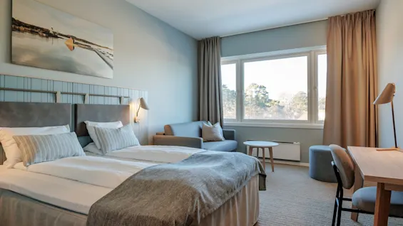 A neatly arranged hotel room features a double bed with gray bedding, a small desk, and a seating area by a large window. A landscape painting decorates the wall.