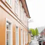 A two-story orange wooden building with white windows rests along a wet, narrow street with parked cars. Signs read "HAMMER" and "HOTEL". Trees and mountains are visible in the distance.