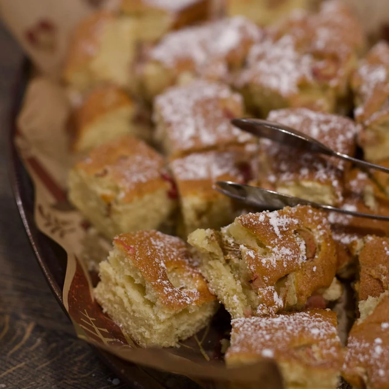 Sliced cake dusted with powdered sugar, tongs resting on top, in a paper-lined tray on a wooden table.