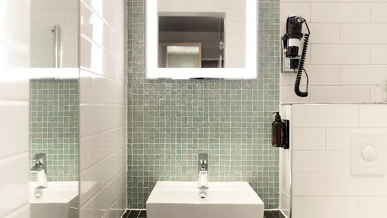 A stylish bathroom at Home Hotel Grand Bodø with a contemporary square sink, a bright, illuminated mirror, and green mosaic tiles.
