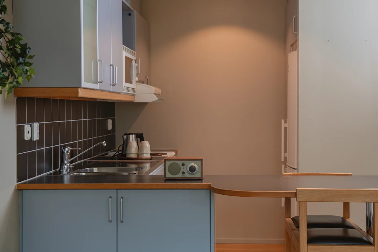 Ett litet kök med ljusblå skåp, en diskho och en bänkskiva med en radio och två vattenkokare. Ett matbord med stolar är delvis synligt. Beige väggar omger.