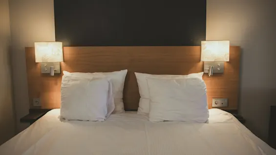 A double bed with white pillows and sheets is flanked by two wall-mounted lamps, casting a warm glow, in a minimalist room with a dark accent wall.