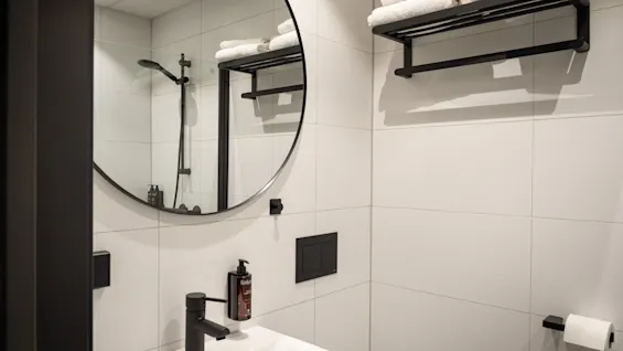 A clean and modern bathroom featuring a white tiled wall, a round mirror, a sink with a black faucet, and fresh towels on a rack.