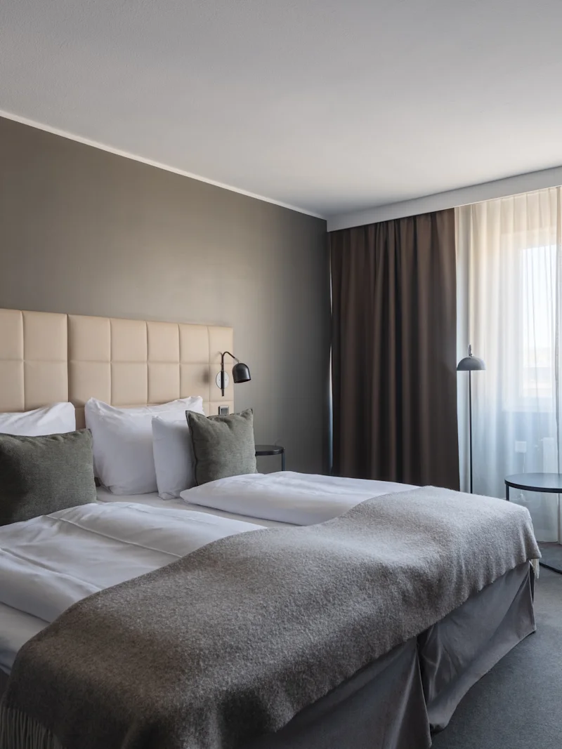 A modern hotel room with a large bed, a comfortable armchair, and a window with sheer and blackout curtains.