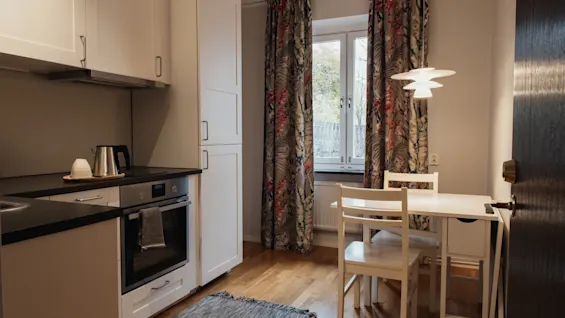 A cozy kitchen and dining area at Home Hotel Kung Oscar with white cabinets, a small table, and patterned curtains.