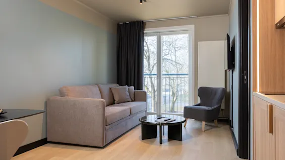 Ett mysigt vardagsrum med soffa, fåtölj och soffbord, med en balkong och utsikt över träd.