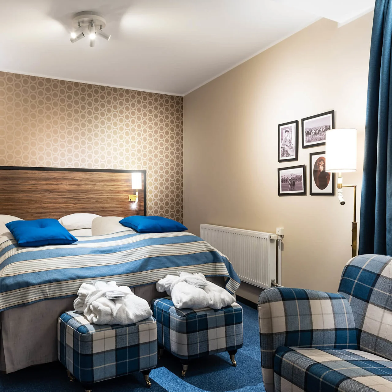 Ett välbäddat säng med blått och vitt lakan i ett välbelyst hotellrum, omgiven av två rutiga fåtöljer och väggkonst.