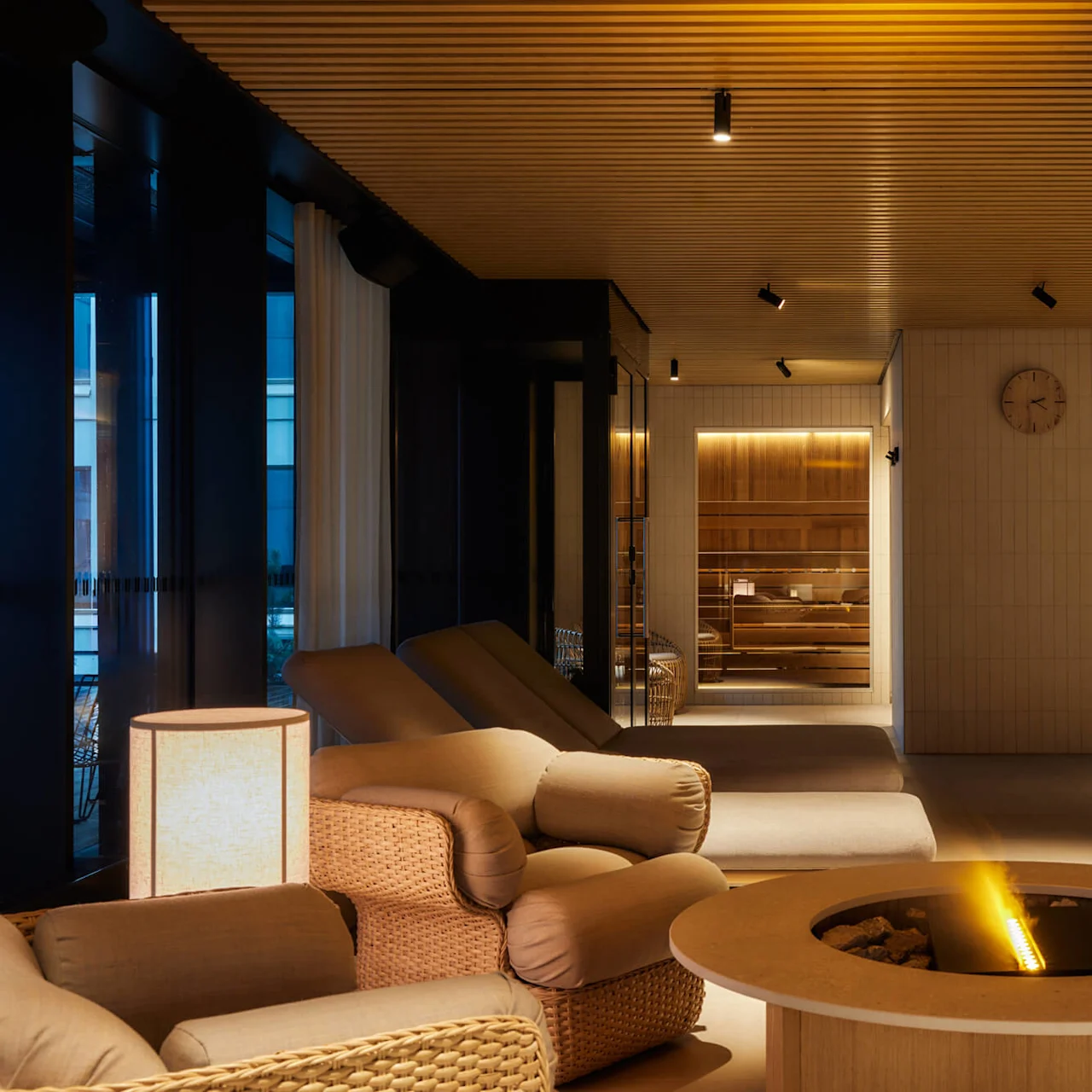 Cozy, cushioned chairs surround a round fireplace with a flickering flame in a warmly lit lounge. Wood paneling and a wall clock enhance the room's calming ambiance.